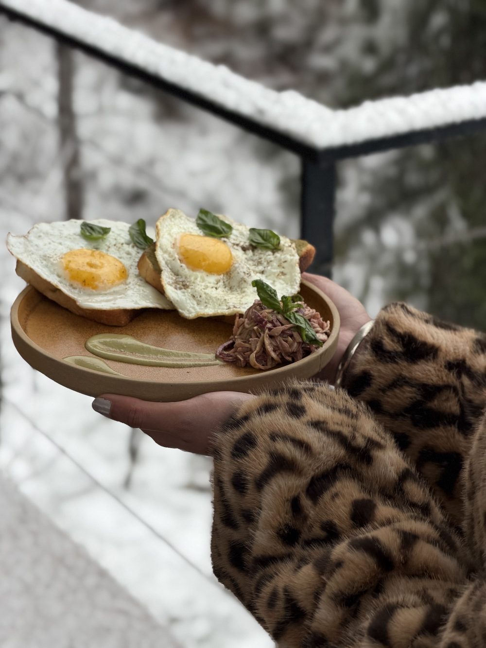 A plate of fried eggs on toast held out over a snowy railing.