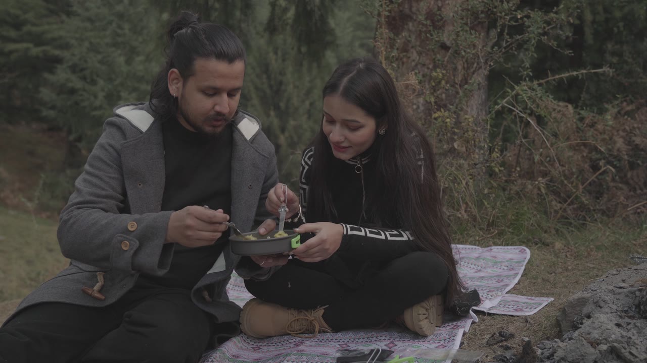 Two guests sharing food on a picnic blanket at the forest edge.