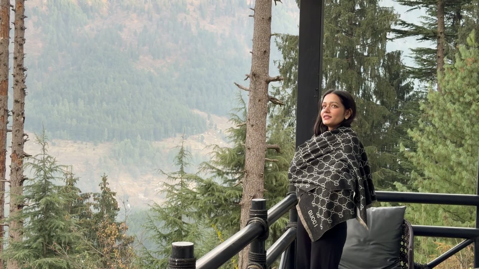 A woman in a shawl on a balcony, looking out over hazy pines.