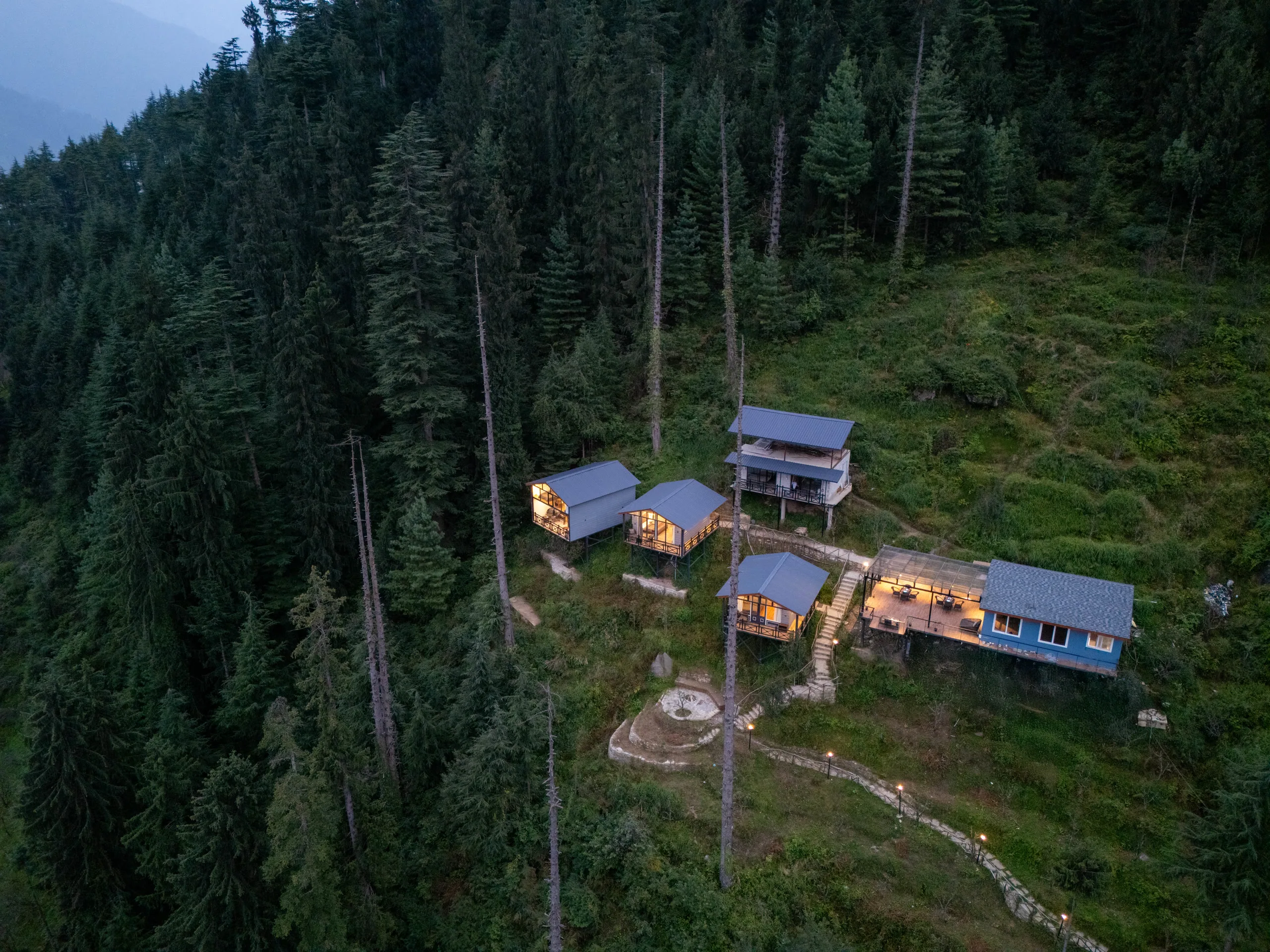 Aerial dusk view of Aarzu Resorts: three lit chalets and the café deck on a forested hillside above the Manali valley.